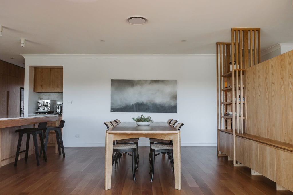Wide view of a dining room wit timber table, rattan chairs and grey artwork with timber cabinetry